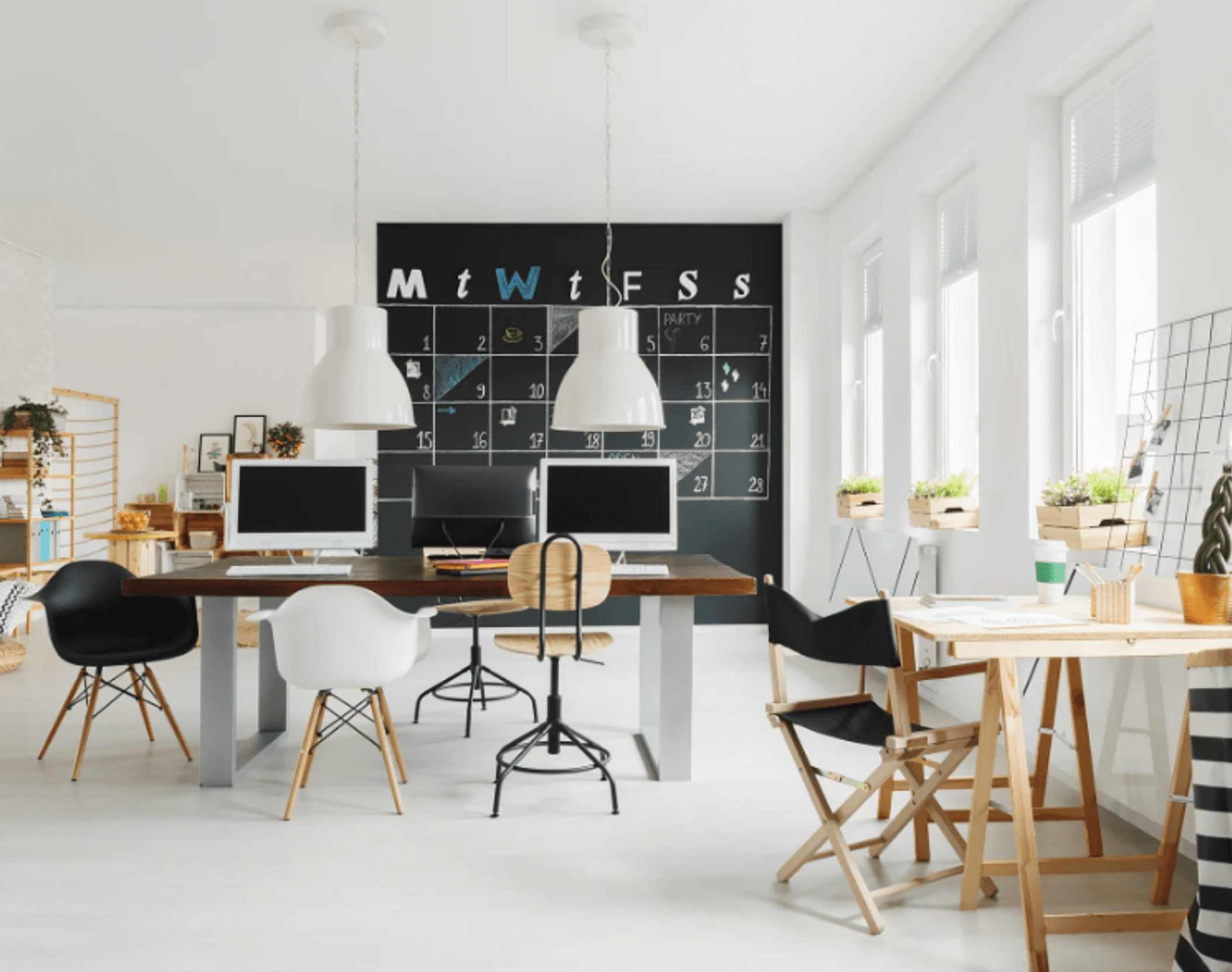 A bright, airy home workspace with a chalkboard wall calendar, pendant lamps, and pale wood furniture